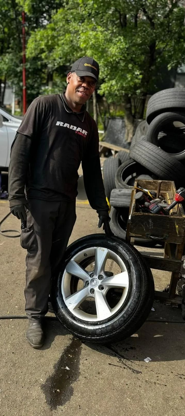 Person in black shirt holding a car tire. Stacks of tires are in the background, with greenery.