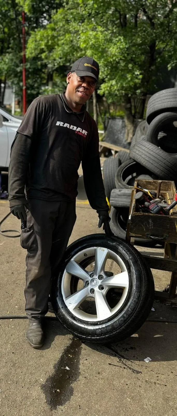 Person in black shirt holding a car tire. Stacks of tires are in the background, with greenery.