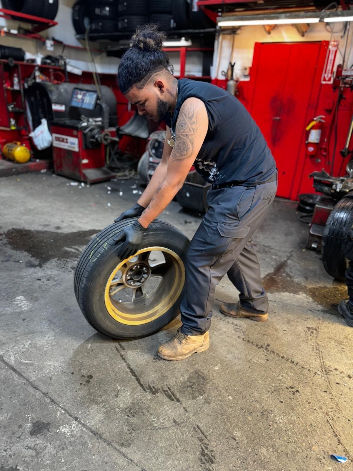 Mechanic in a garage holding a tire. He's wearing black gloves, a black shirt, and gray pants.