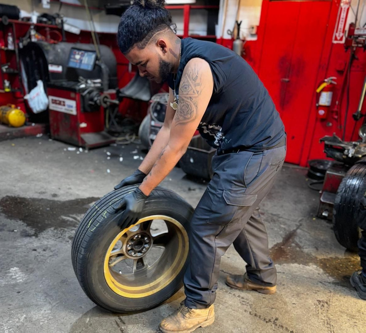 Mechanic handling a tire, inside a garage. He is wearing gloves and a black shirt.