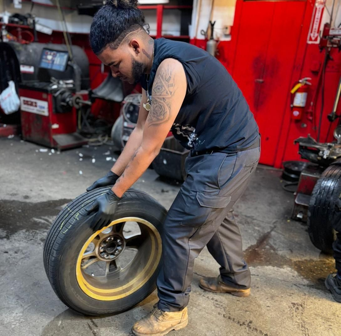 Mechanic handling a tire, inside a garage. He is wearing gloves and a black shirt.