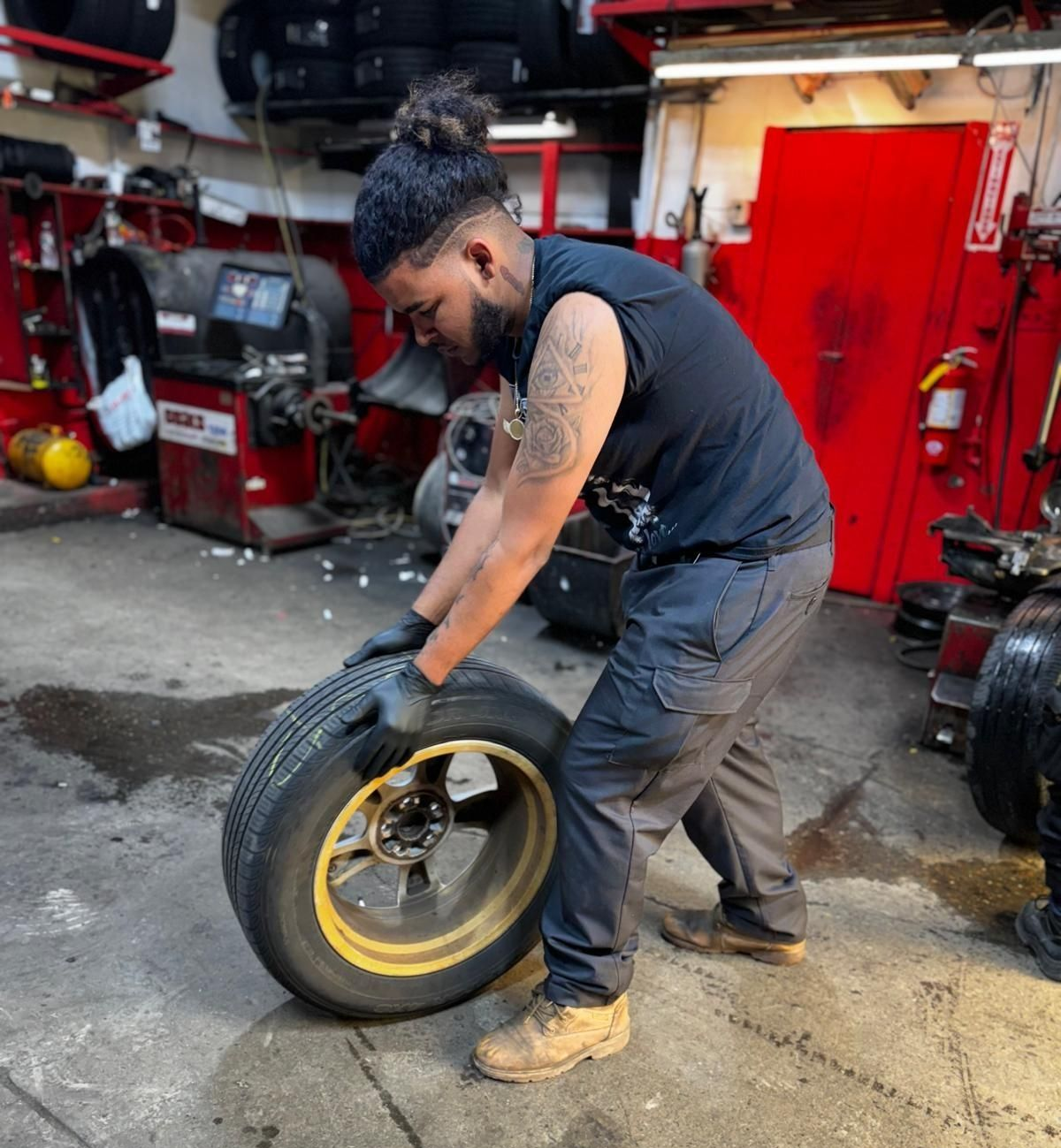 Mechanic in a garage holding a tire. He's wearing black gloves, a black shirt, and gray pants.