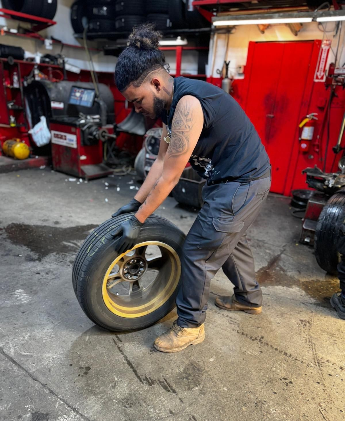Mechanic in a garage holding a tire. He's wearing black gloves, a black shirt, and gray pants.