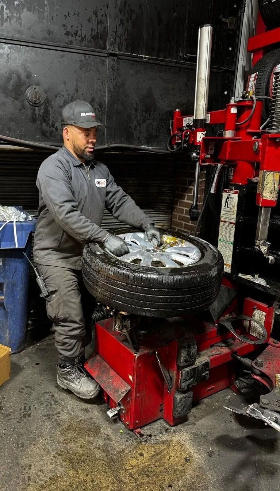 Mechanic changing a tire on a red machine in a shop. He wears a cap, gray work suit, gloves.