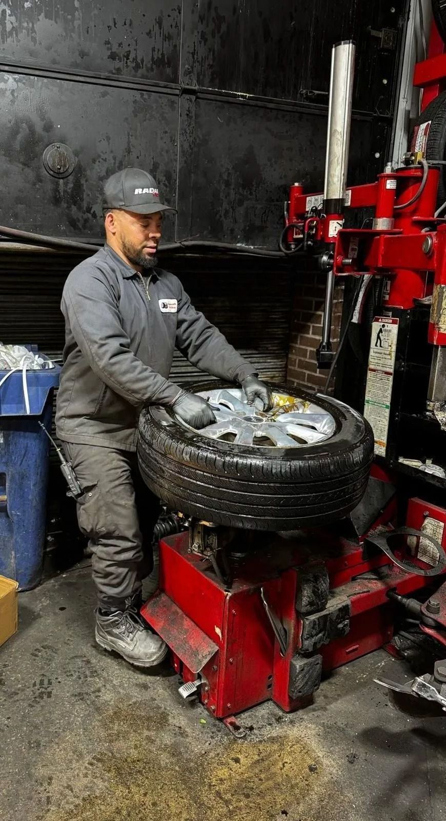 Mechanic changing a tire on a red machine in a shop. He wears a cap, gray work suit, gloves.