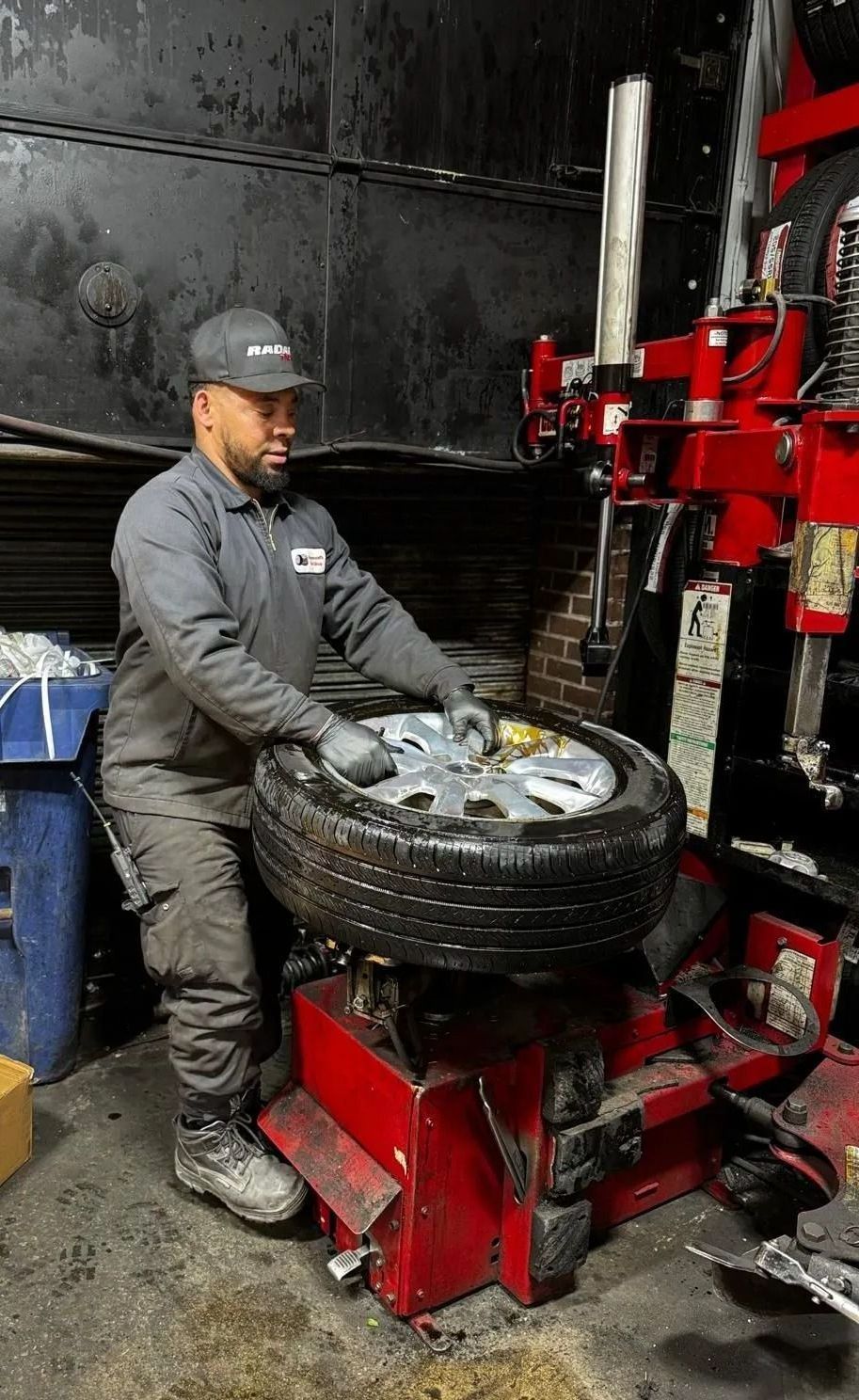 Mechanic changing a tire on a red machine in a shop. He wears a cap, gray work suit, gloves.