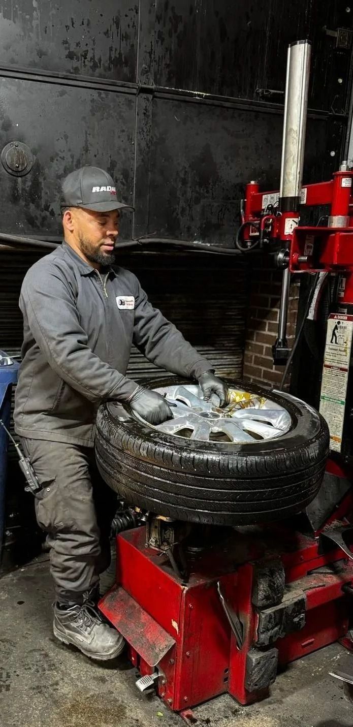Mechanic changing a tire on a red machine in a shop. He wears a cap, gray work suit, gloves.