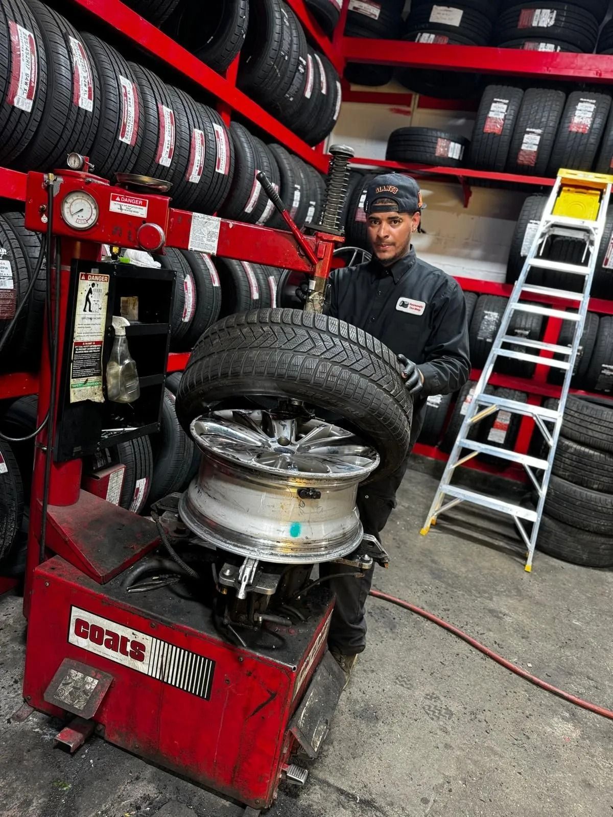 Man at tire shop changing a tire on a red machine; surrounded by tires, standing next to a ladder.
