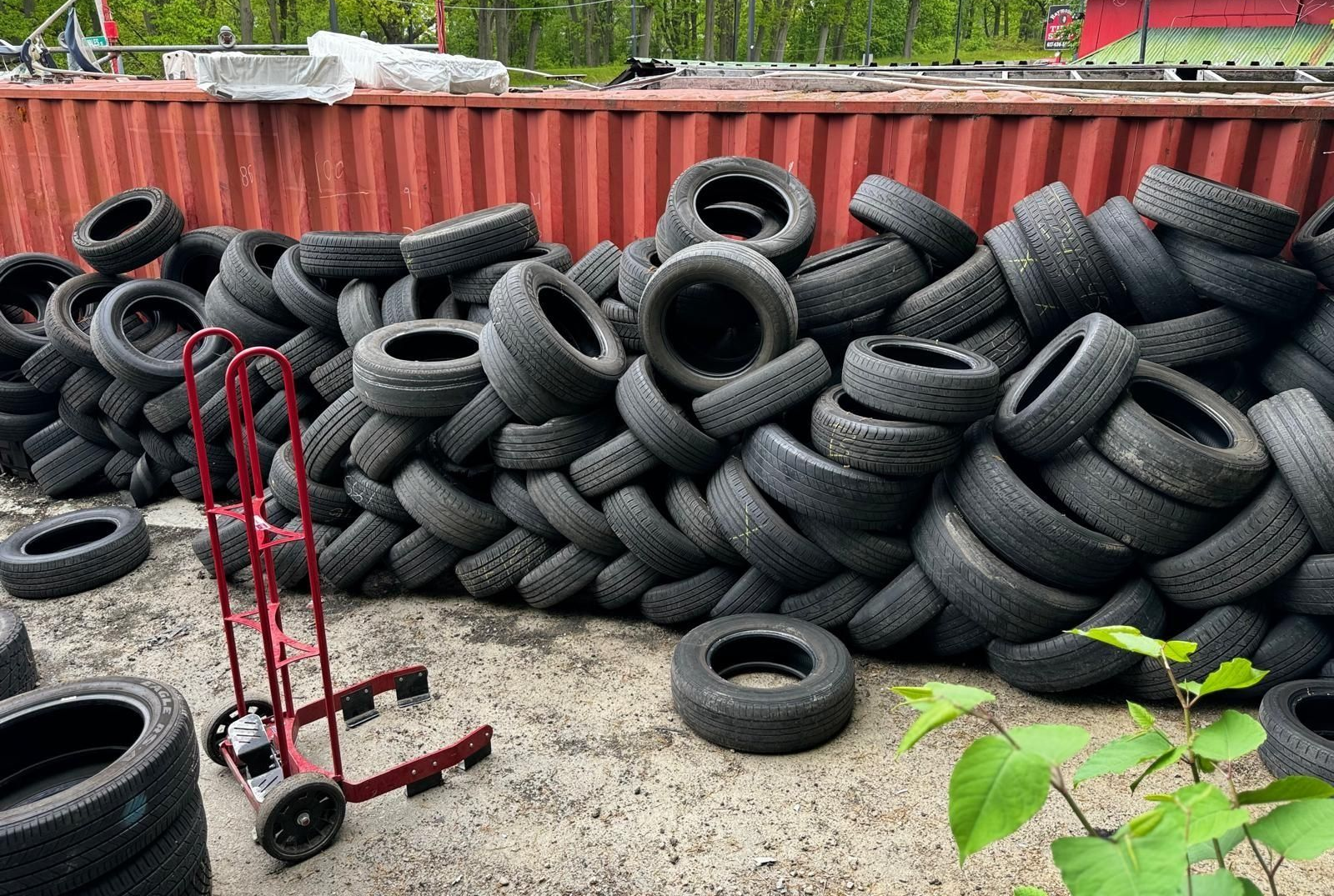 Pile of black tires next to a red hand truck; outdoors by a red container and green trees.