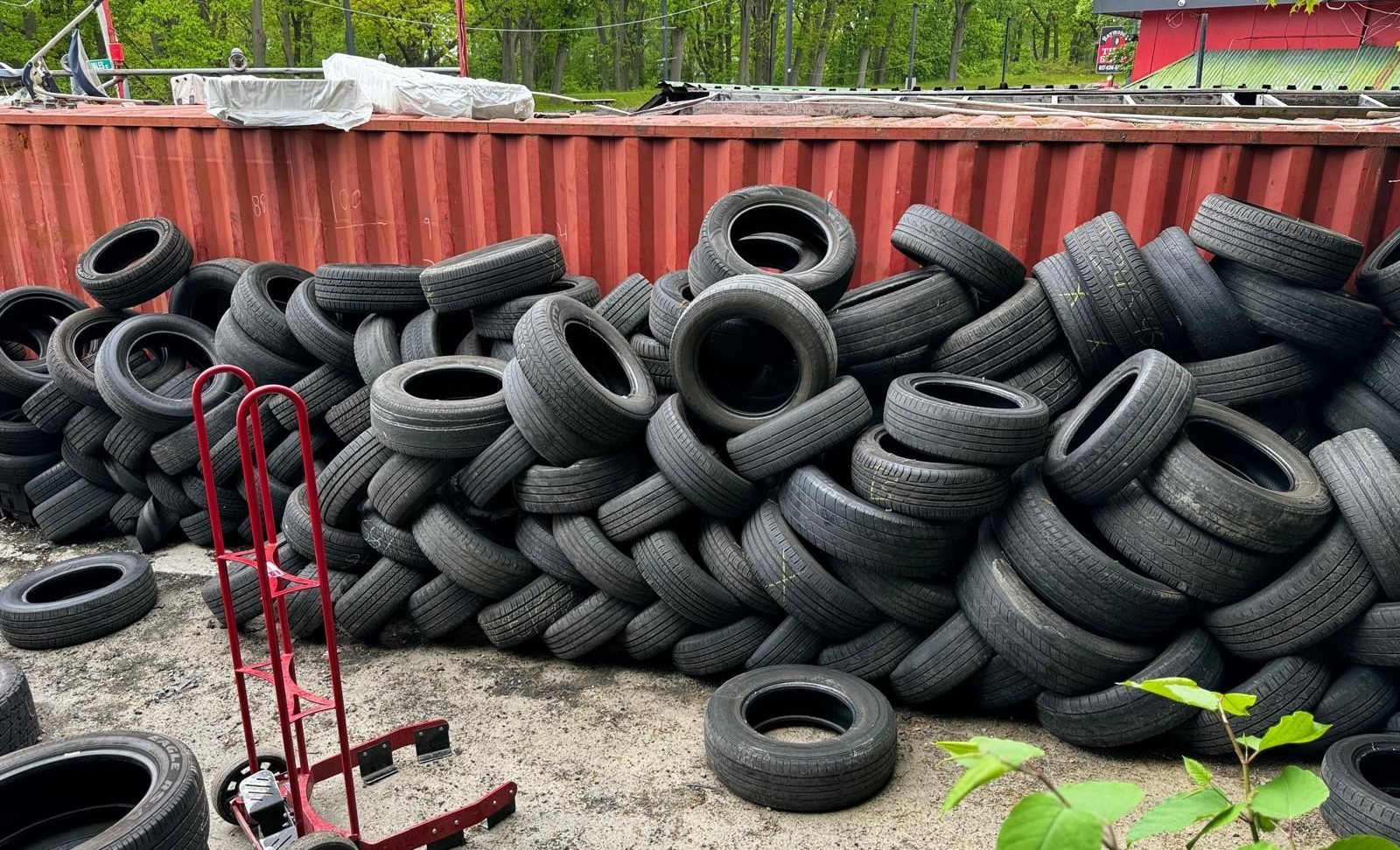 Pile of used tires against a red container, with a red hand truck and foliage in the foreground.