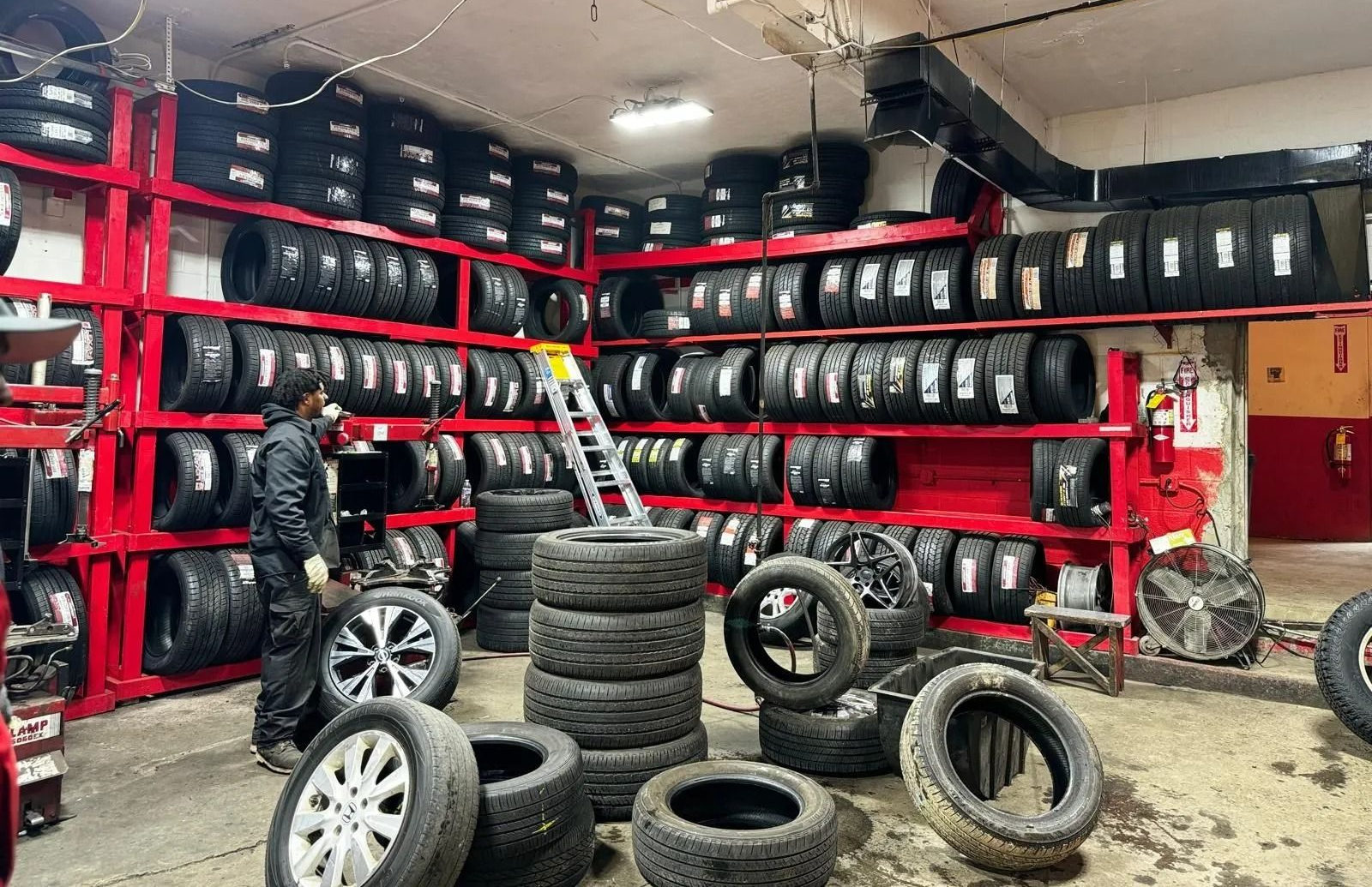 A mechanic in a shop with many tires on red shelves. Some tires on the floor, ladder present.