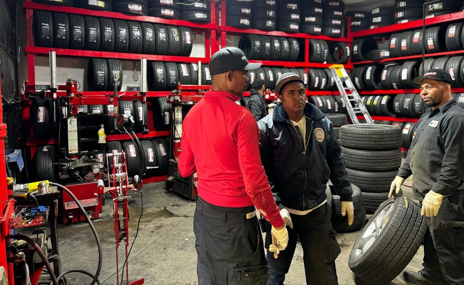 Three men in a tire shop. One holds a tire, others converse near equipment and shelves of tires.