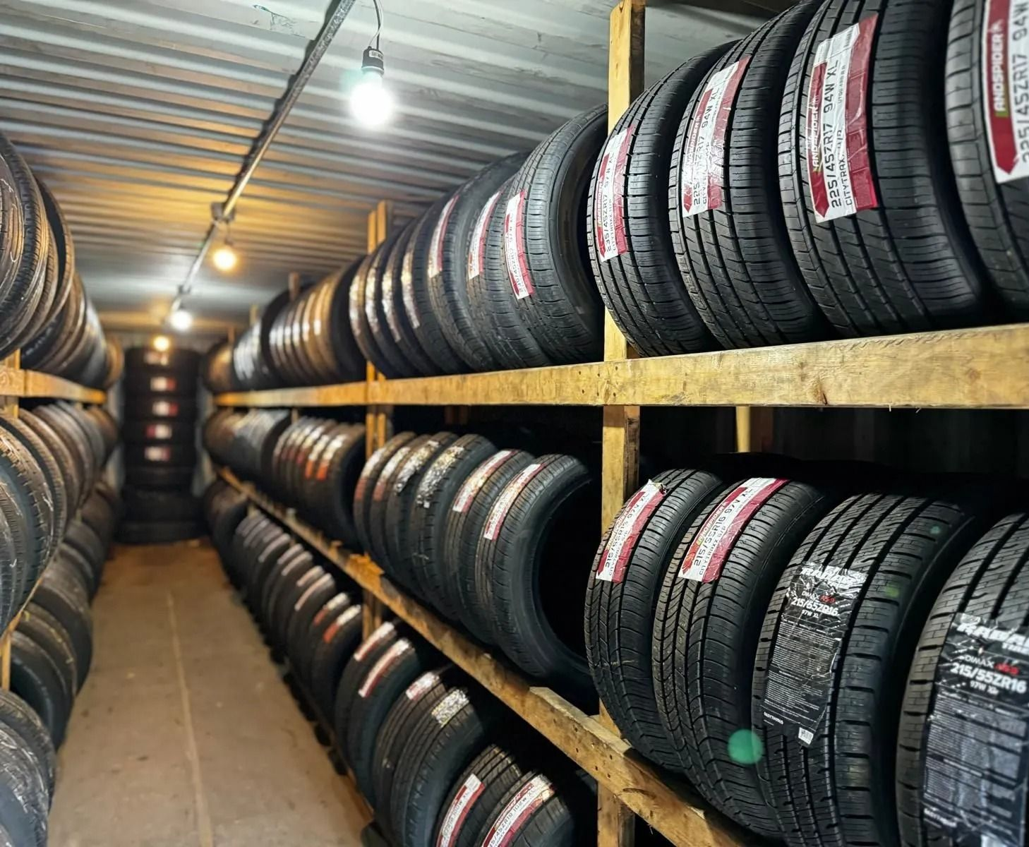 Tires stacked on wooden shelves in a storage room, lit by overhead lights.