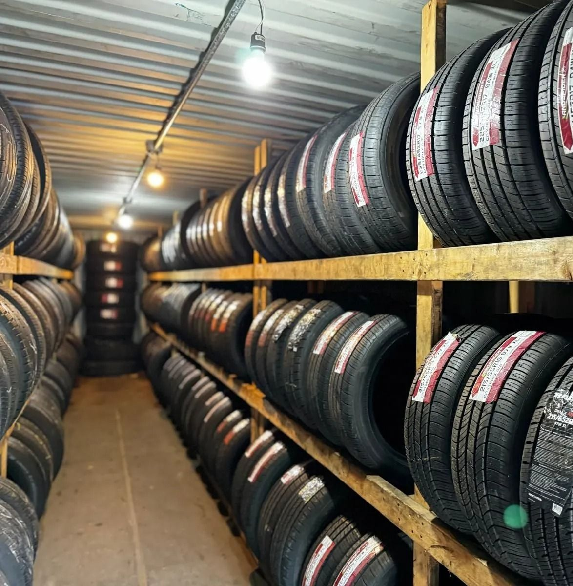 Tires stacked on wooden shelves in a storage room, lit by overhead lights.
