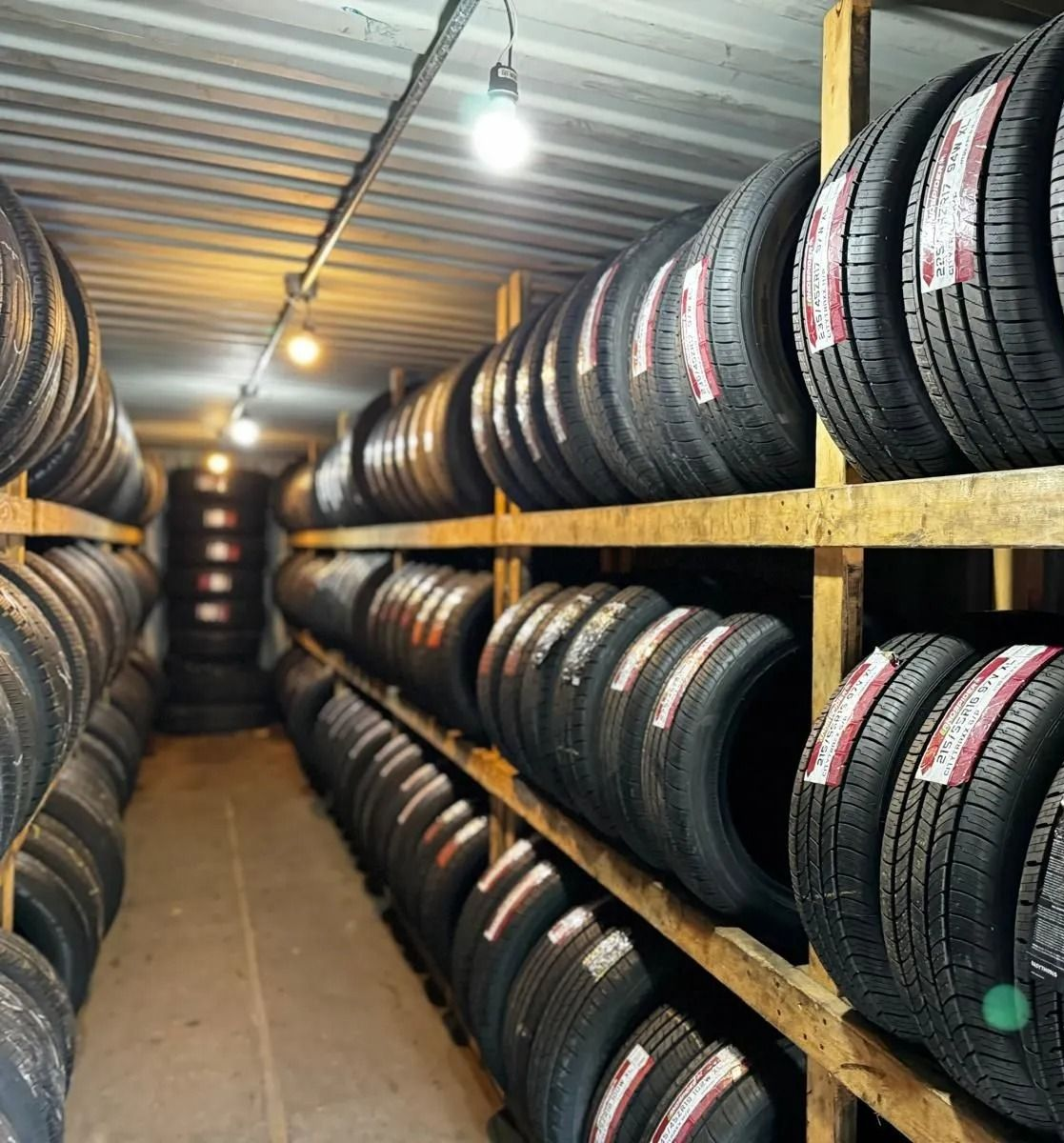 Tires stacked on wooden shelves in a storage room, lit by overhead lights.