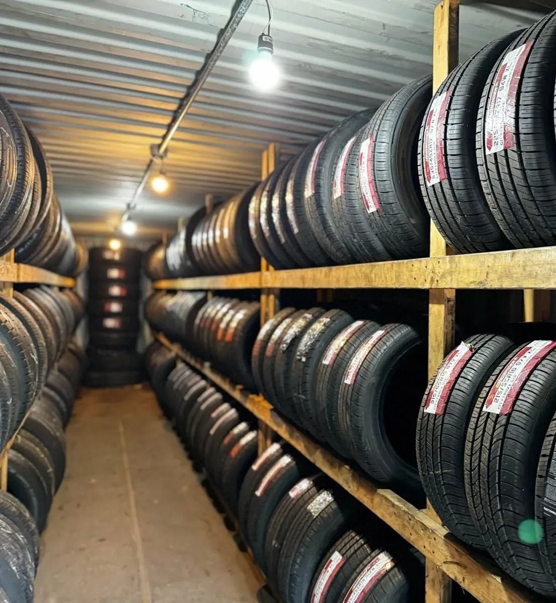 Tires stacked on wooden shelves in a storage room, lit by overhead lights.