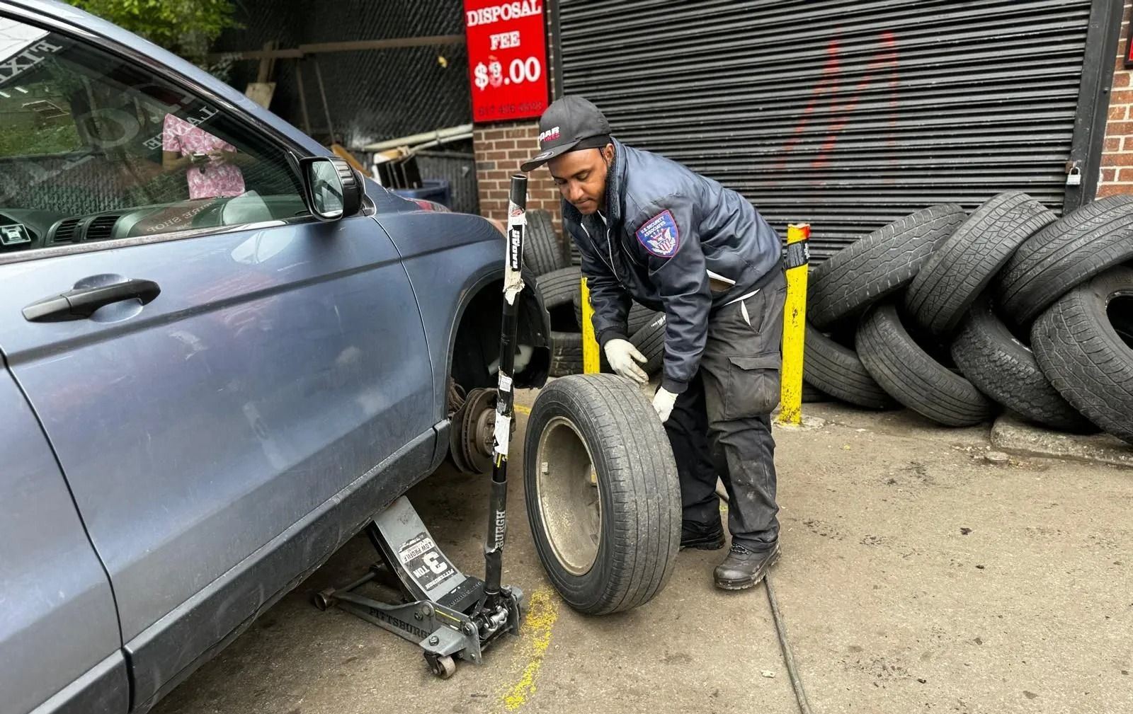 Mechanic changing a tire on a gray car, standing outside a shop with stacked tires.