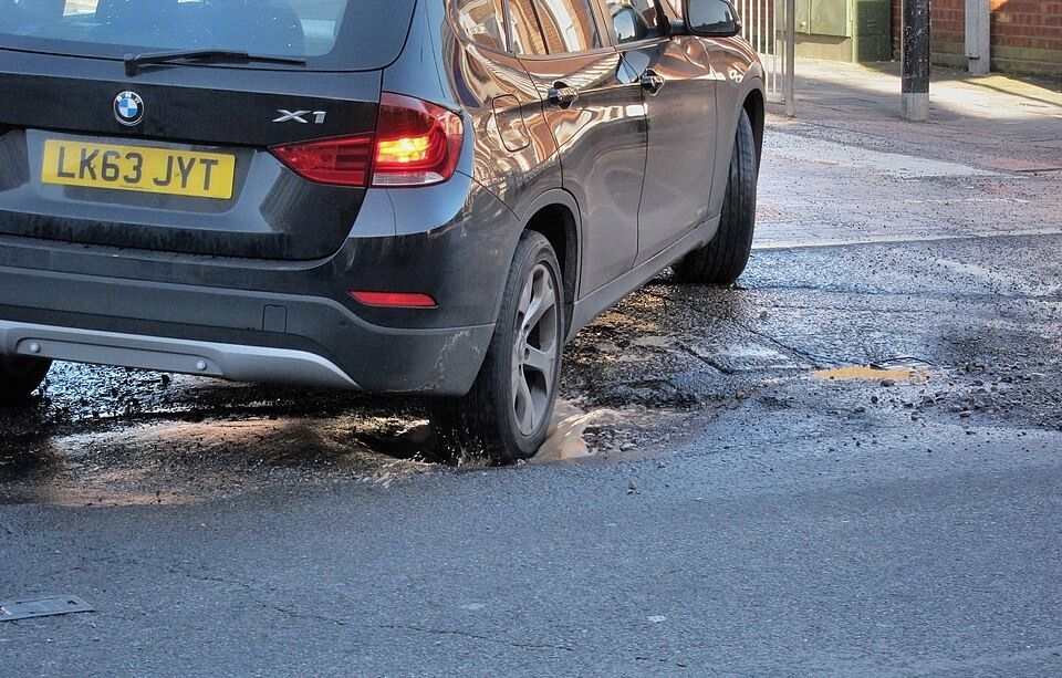 A black BMW X1 SUV driving through a deep, muddy pothole on an urban road.