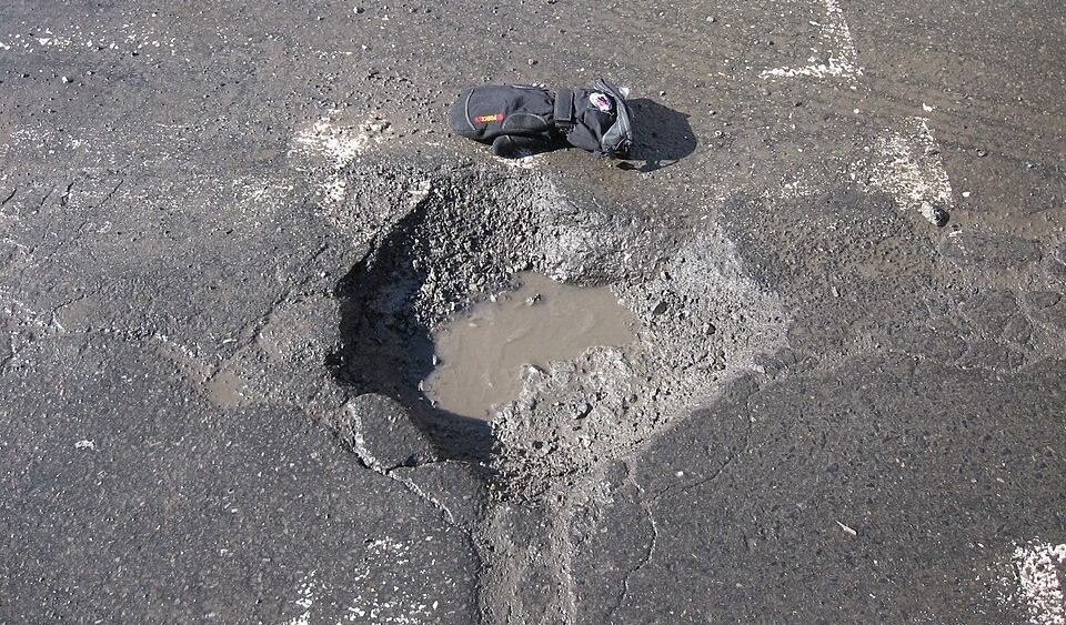 A large, dark pothole in asphalt pavement containing a small puddle, with a black glove placed near the edge for scale.