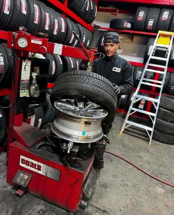 Man changing a tire in a shop, using a tire machine, surrounded by tire racks and a ladder.