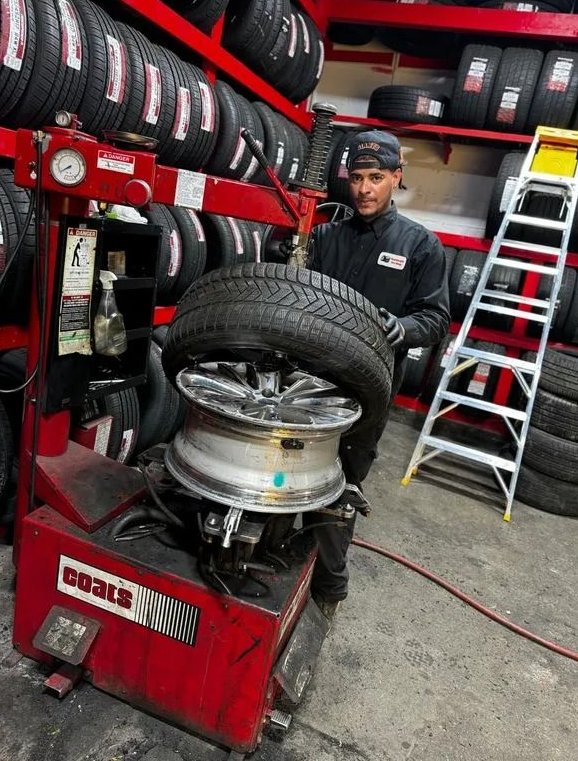 Man changing a tire in a shop, using a tire machine, surrounded by tire racks and a ladder.