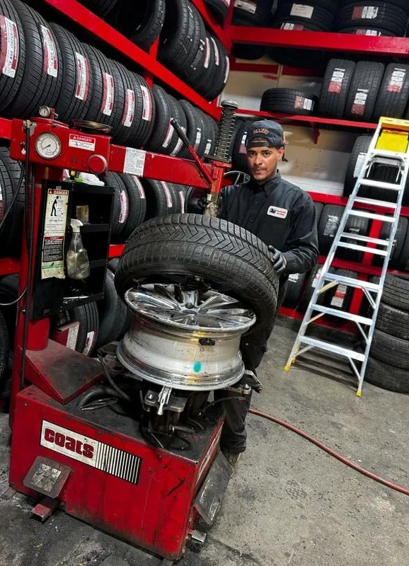 Man changing a tire in a shop, using a tire machine, surrounded by tire racks and a ladder.