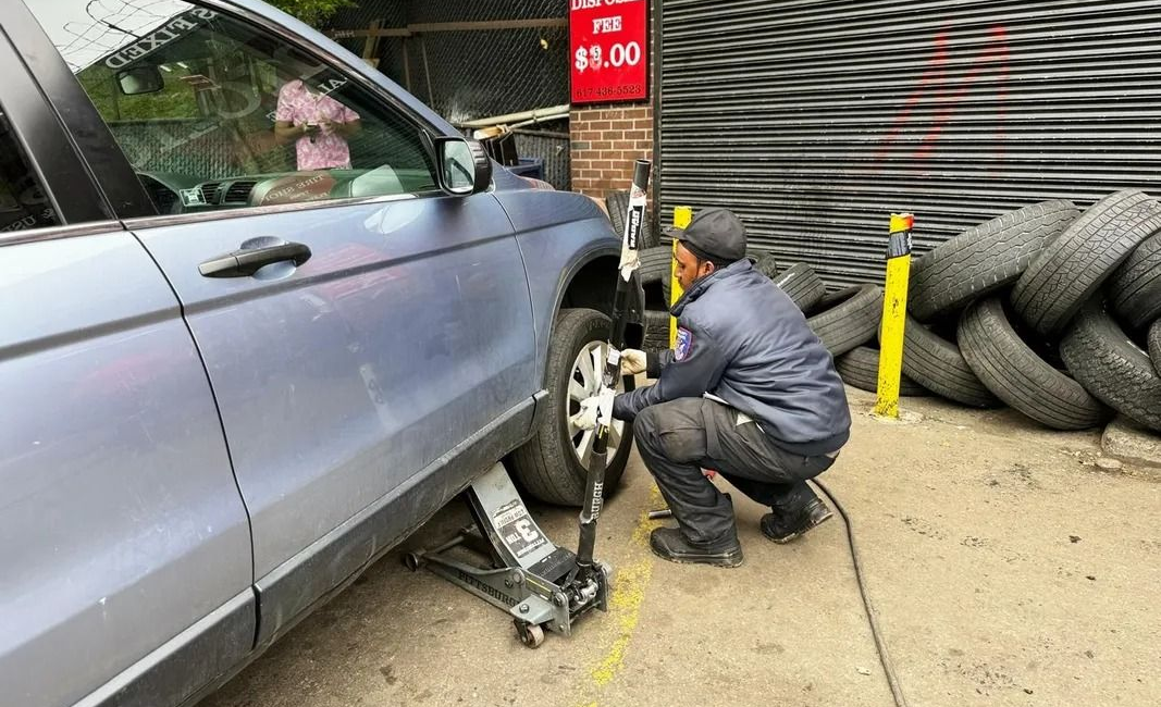 A mechanic aligns the front tire of a silver SUV with tools, outside a tire shop.