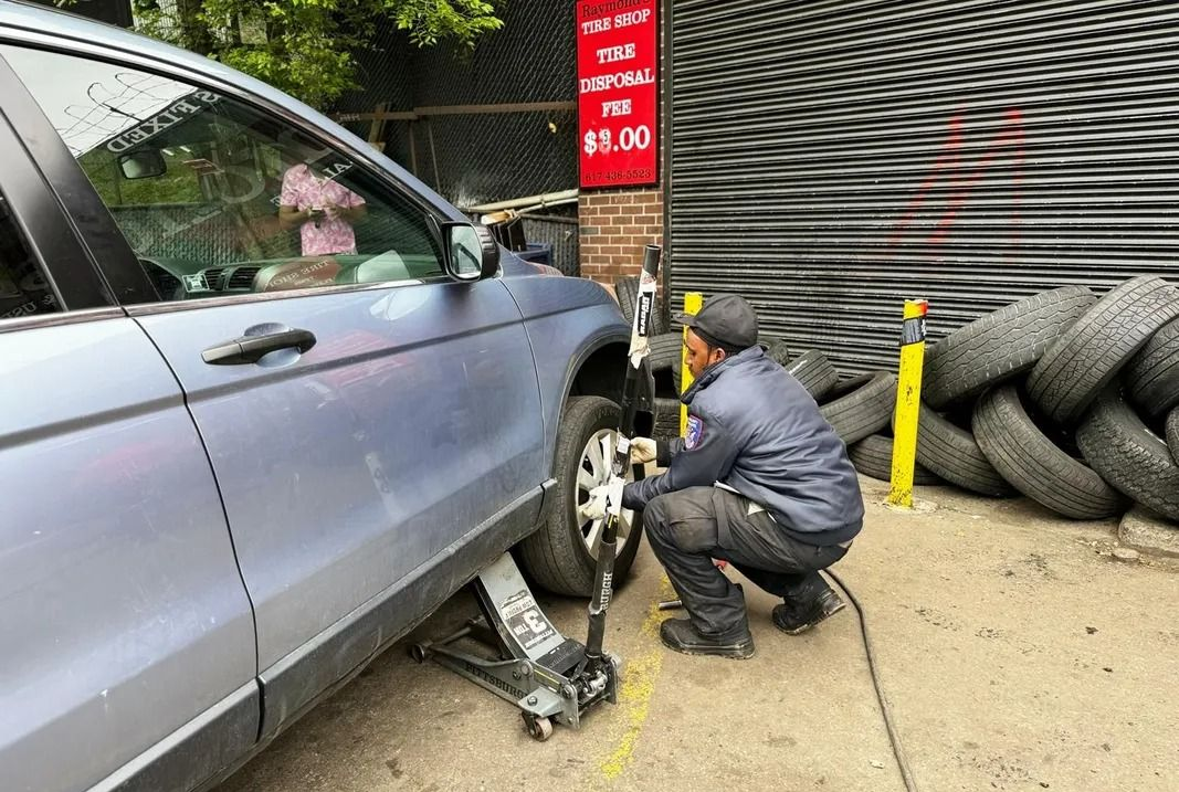 Mechanic changing a car tire at a tire shop. He is kneeling, using a jack. Tires piled nearby.