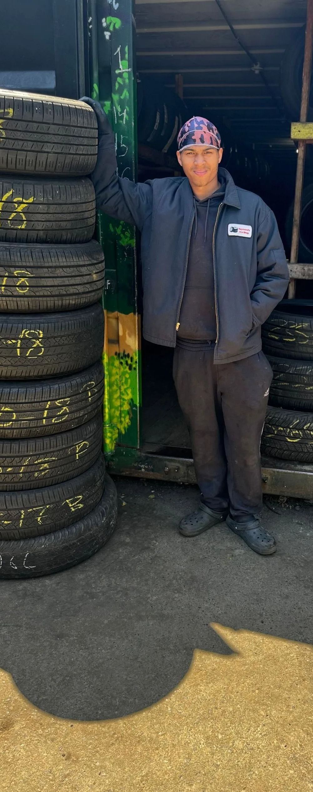 A man stands next to stacked tires, in a garage. He wears a jacket and head covering.