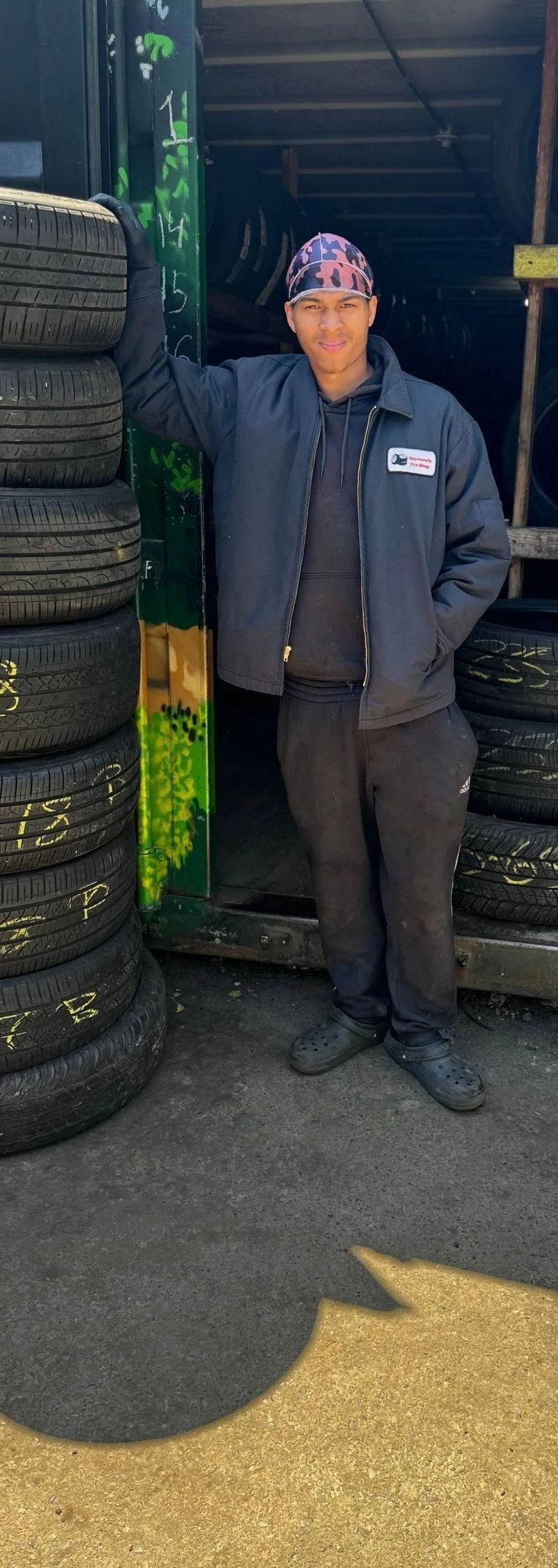 Man in blue jacket and bandana stands near tires, leaning against a stack outside a garage.