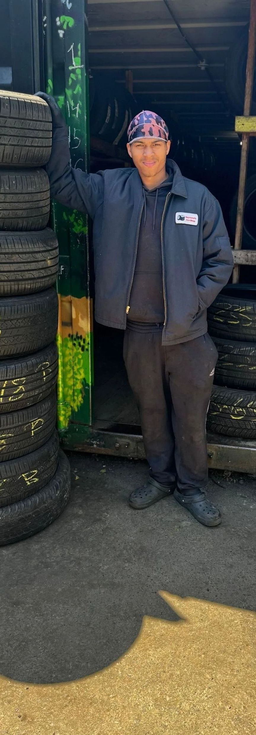 Man in blue jacket and bandana stands near tires, leaning against a stack outside a garage.