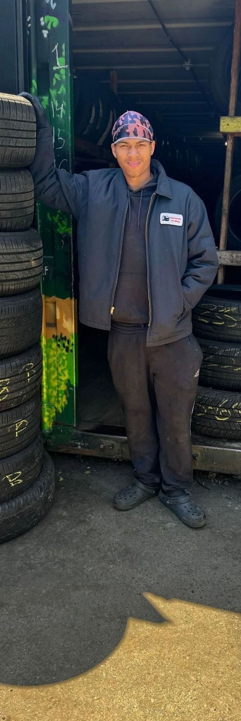 Man in blue jacket and bandana stands near tires, leaning against a stack outside a garage.