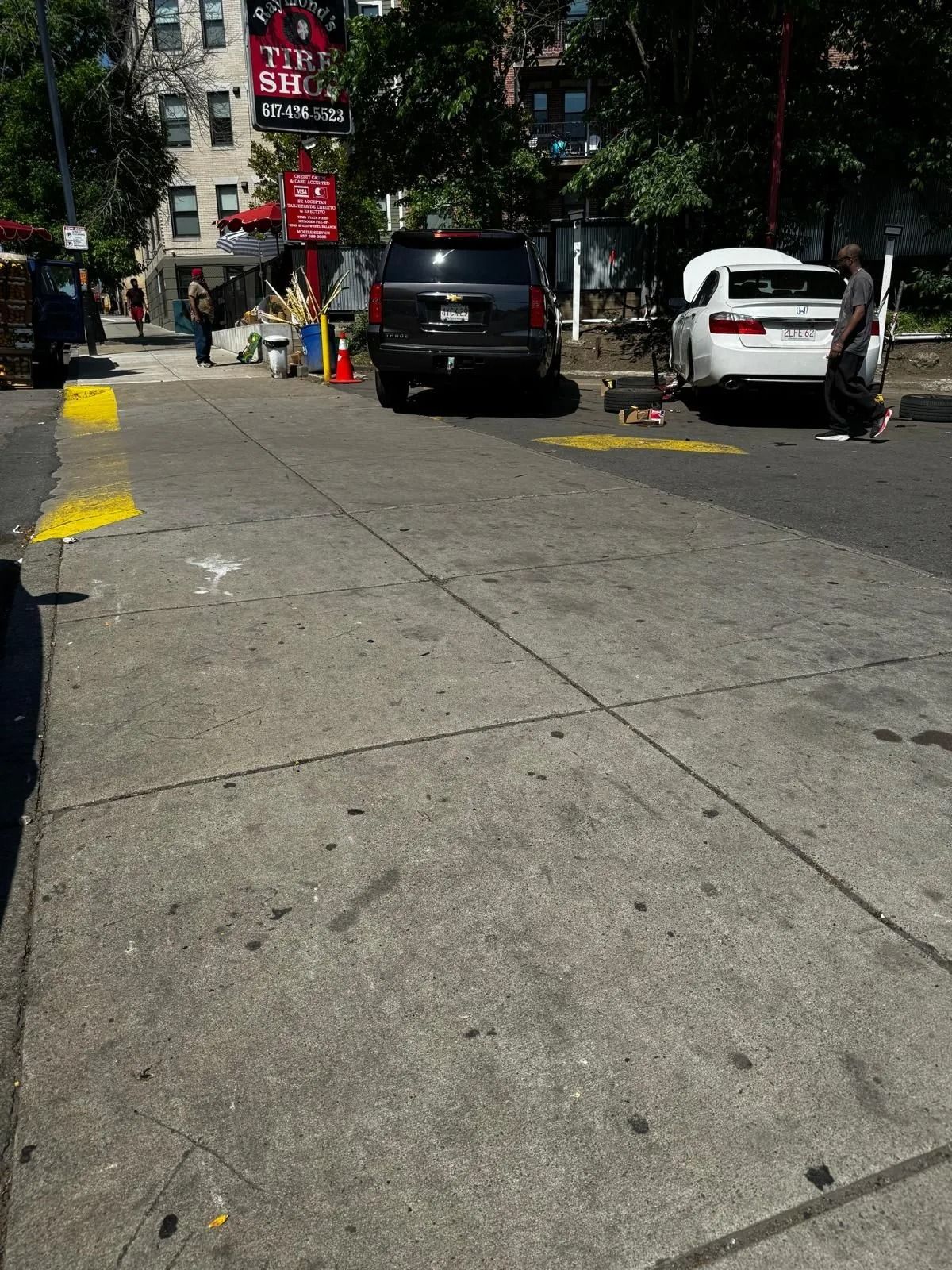 Urban street with parked black and white cars, trees, and a bright sidewalk in sunlight.