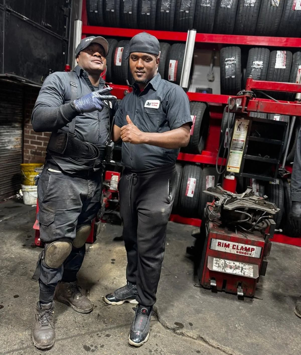 Two mechanics in a tire shop pose; one gives a peace sign, the other a thumbs up. Tires are in the background.
