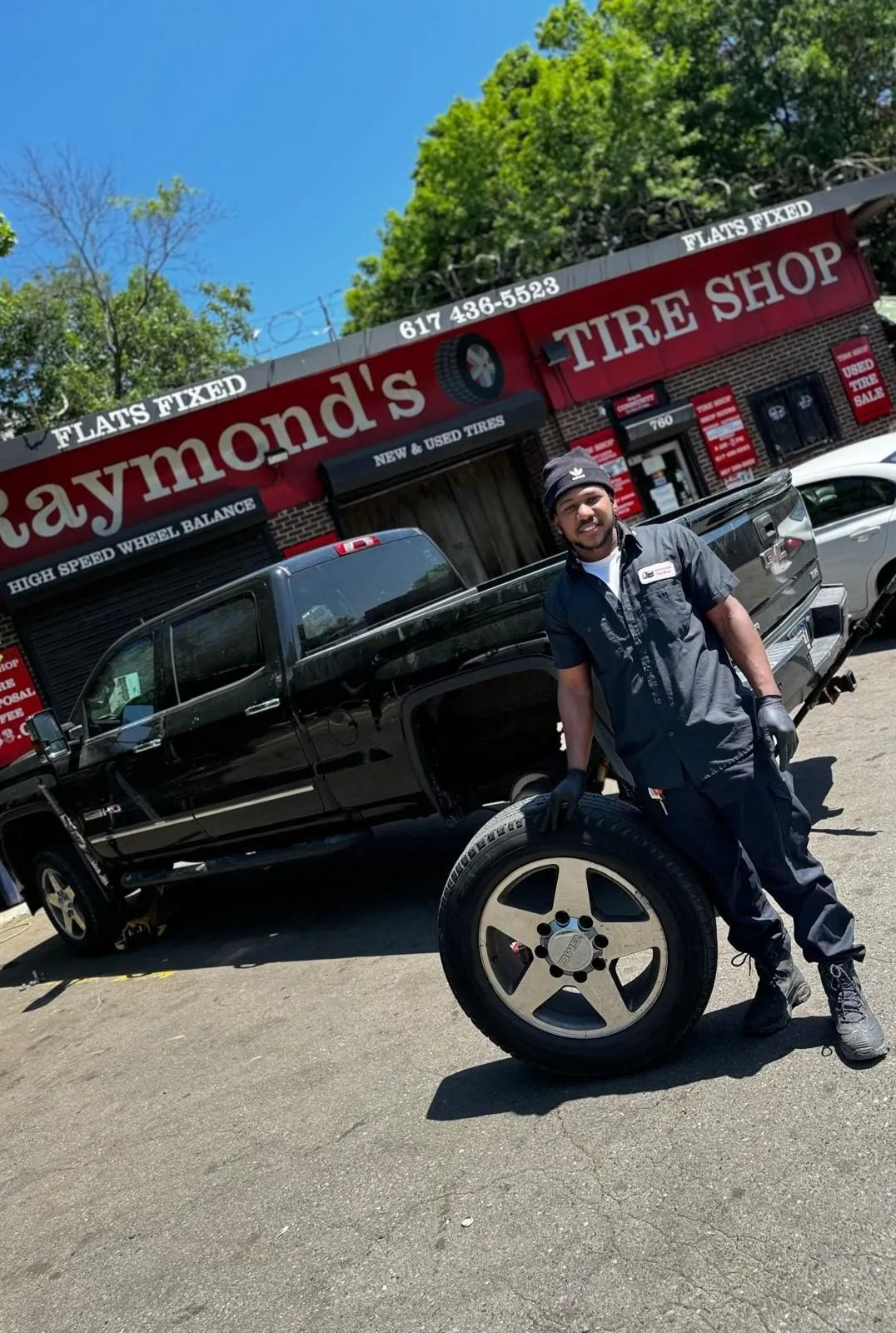 Mechanic poses with a tire in front of Raymond's Tire Shop with a black pickup truck.