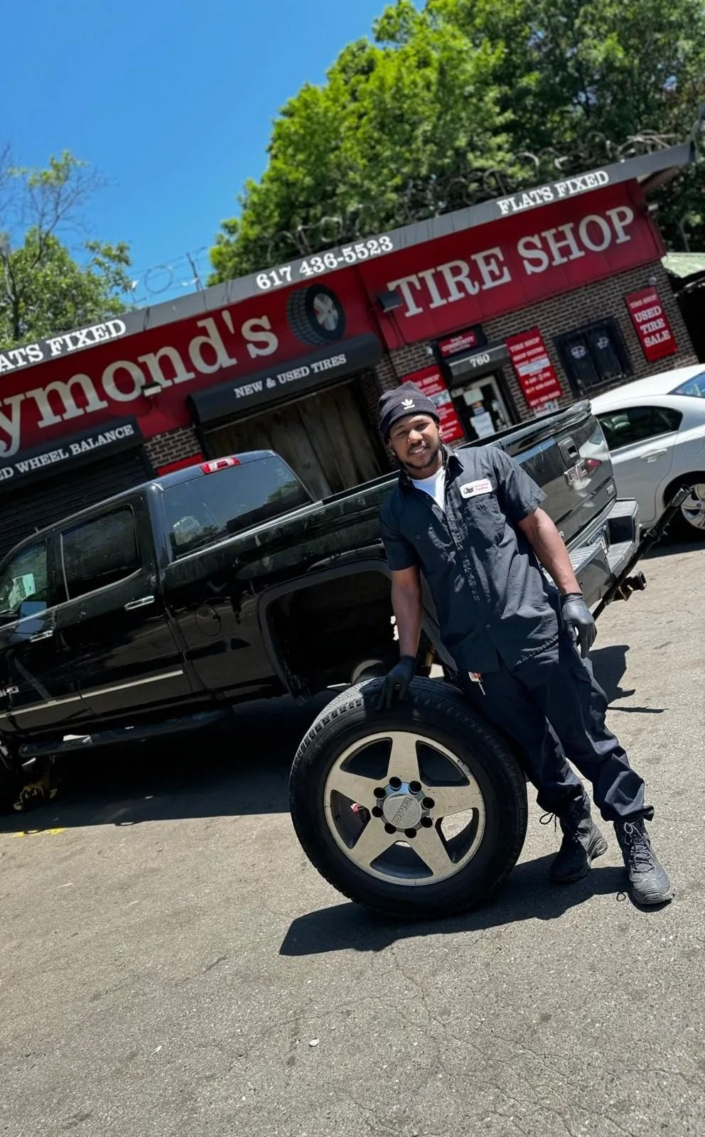 A man in work clothes holds a tire, posing beside a black truck in front of Raymond's Tire Shop.
