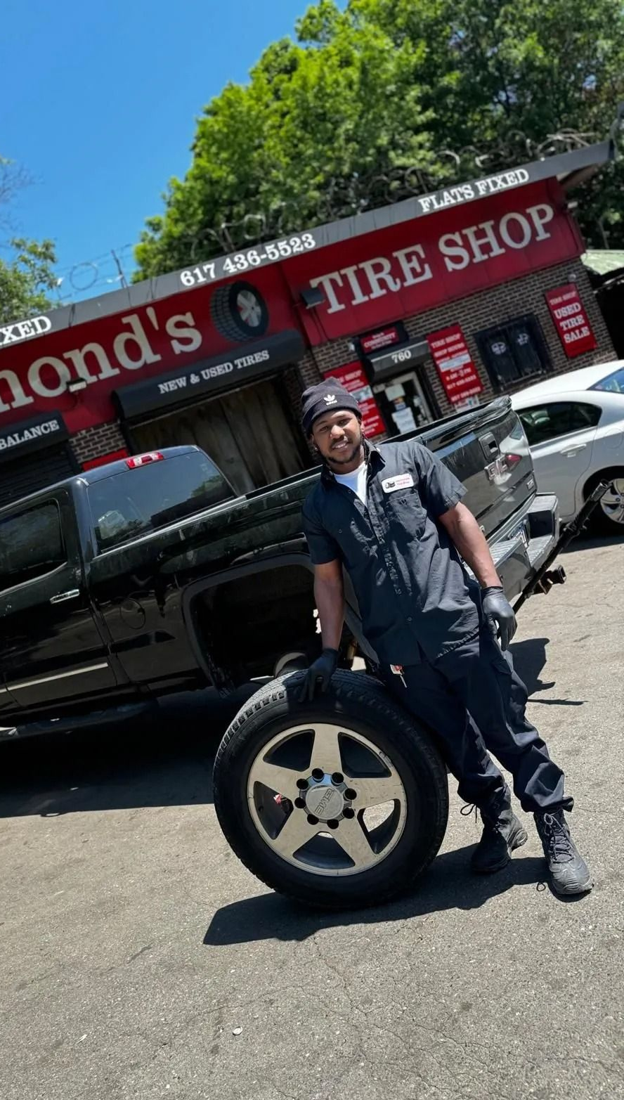 A man in work clothes holds a tire, posing beside a black truck in front of Raymond's Tire Shop.