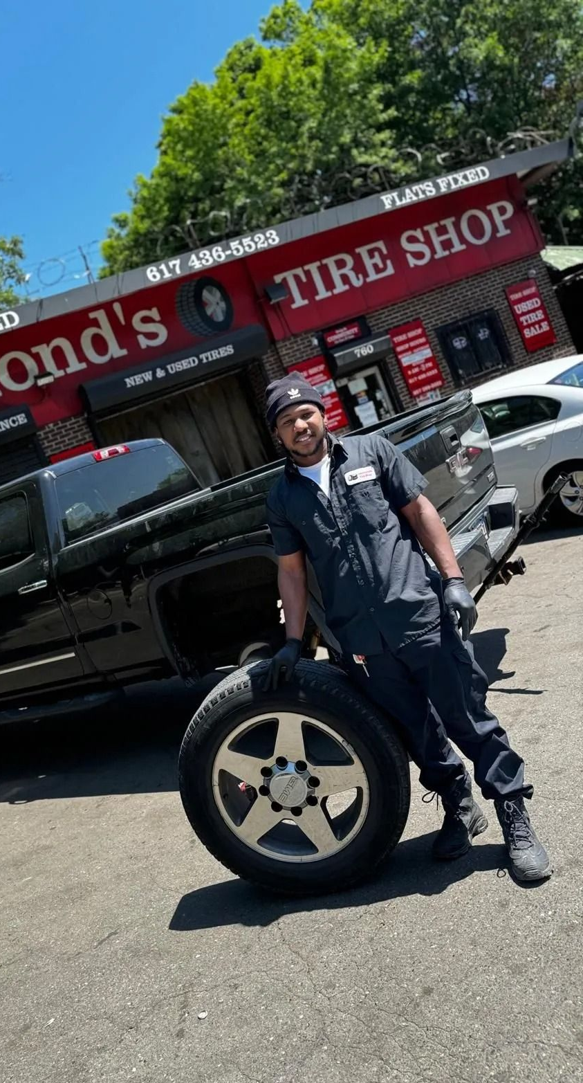 A man in work clothes holds a tire, posing beside a black truck in front of Raymond's Tire Shop.