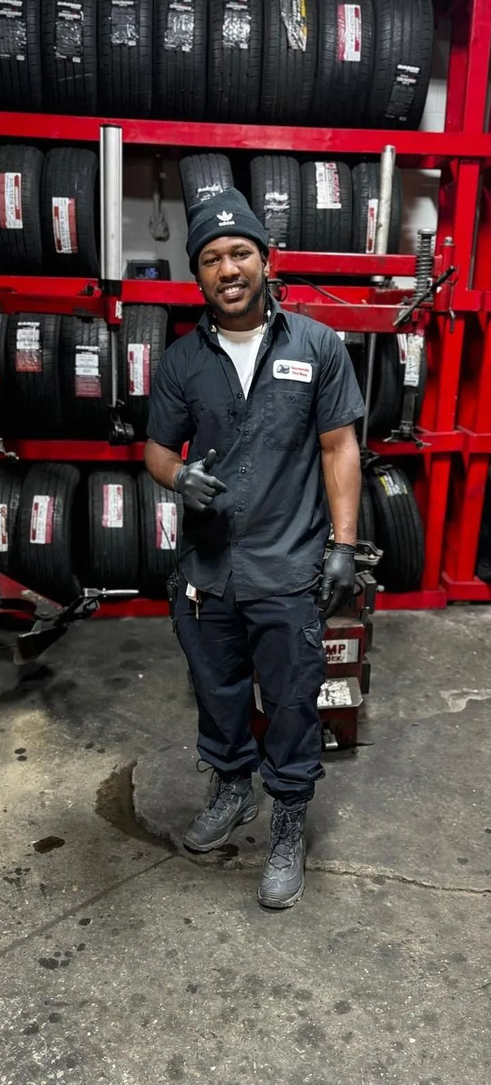 Man in a mechanic uniform smiles in a tire shop; tires on shelves in the background.