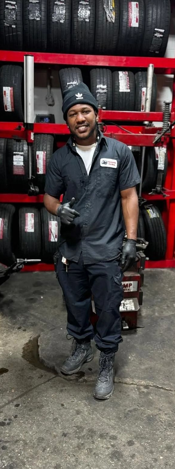 Man in a mechanic uniform smiles in a tire shop; tires on shelves in the background.
