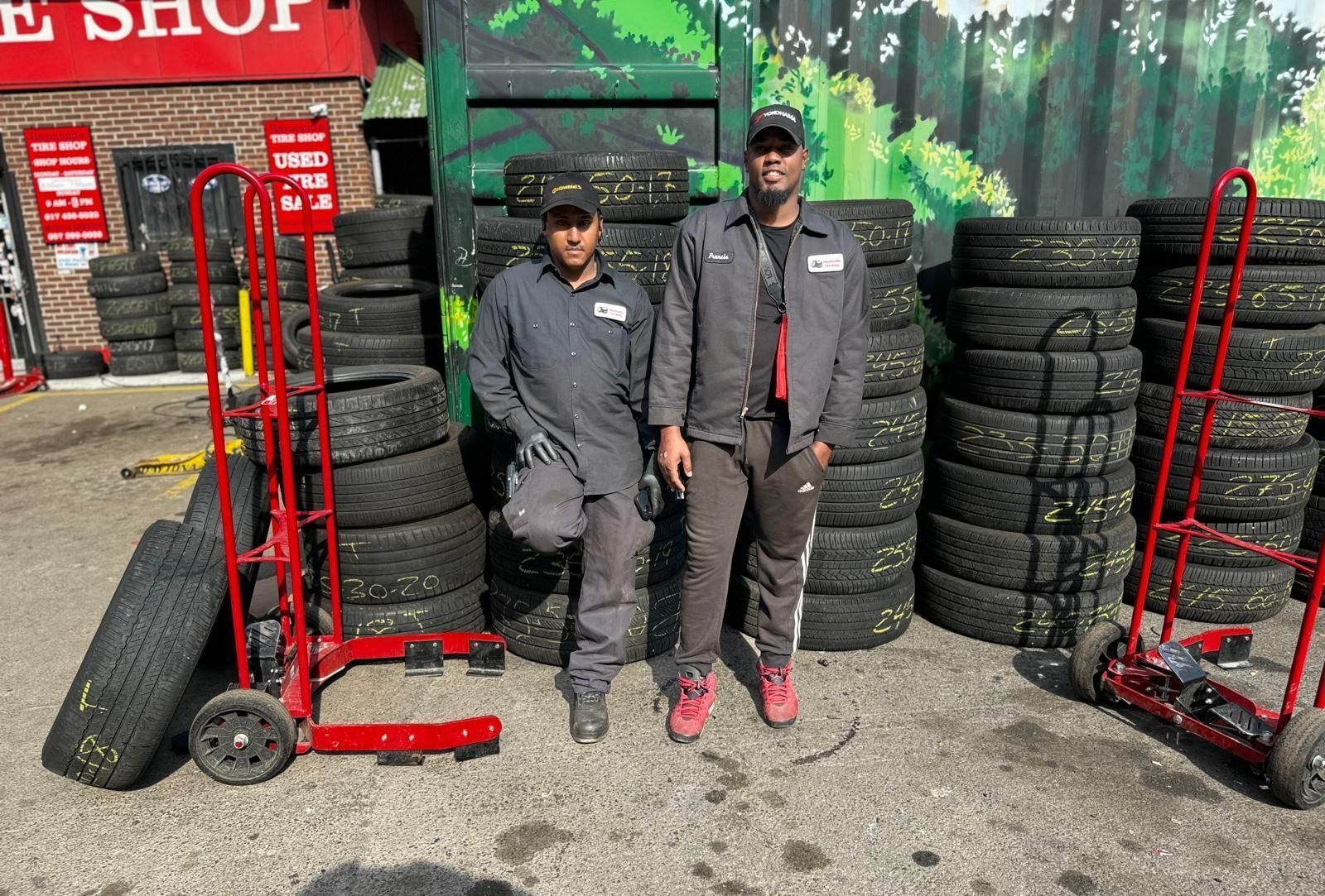 Two men stand by stacks of tires outside a tire shop, with red hand trucks on either side.