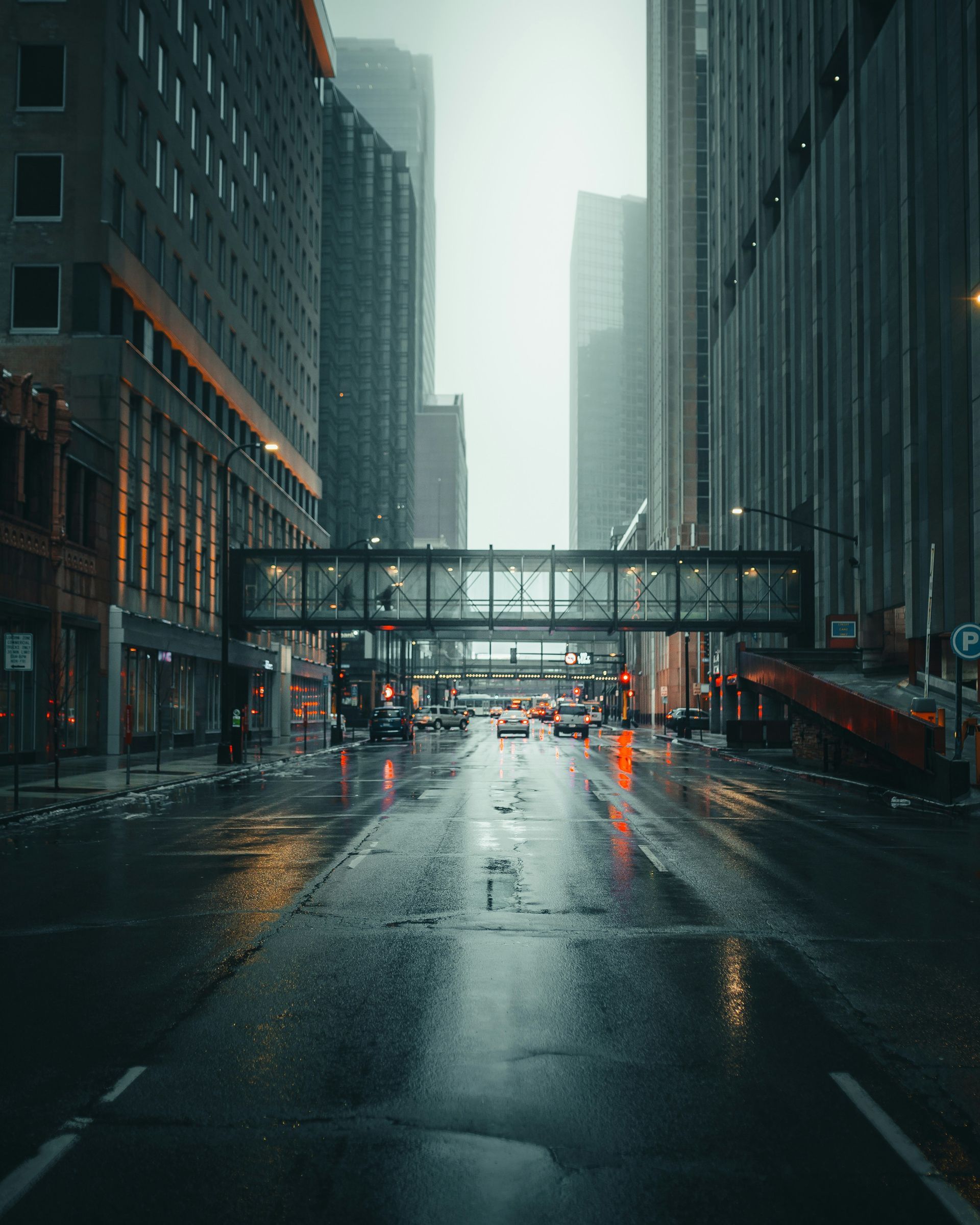 Rainy city street between tall skyscrapers with wet pavement and a pedestrian bridge overhead