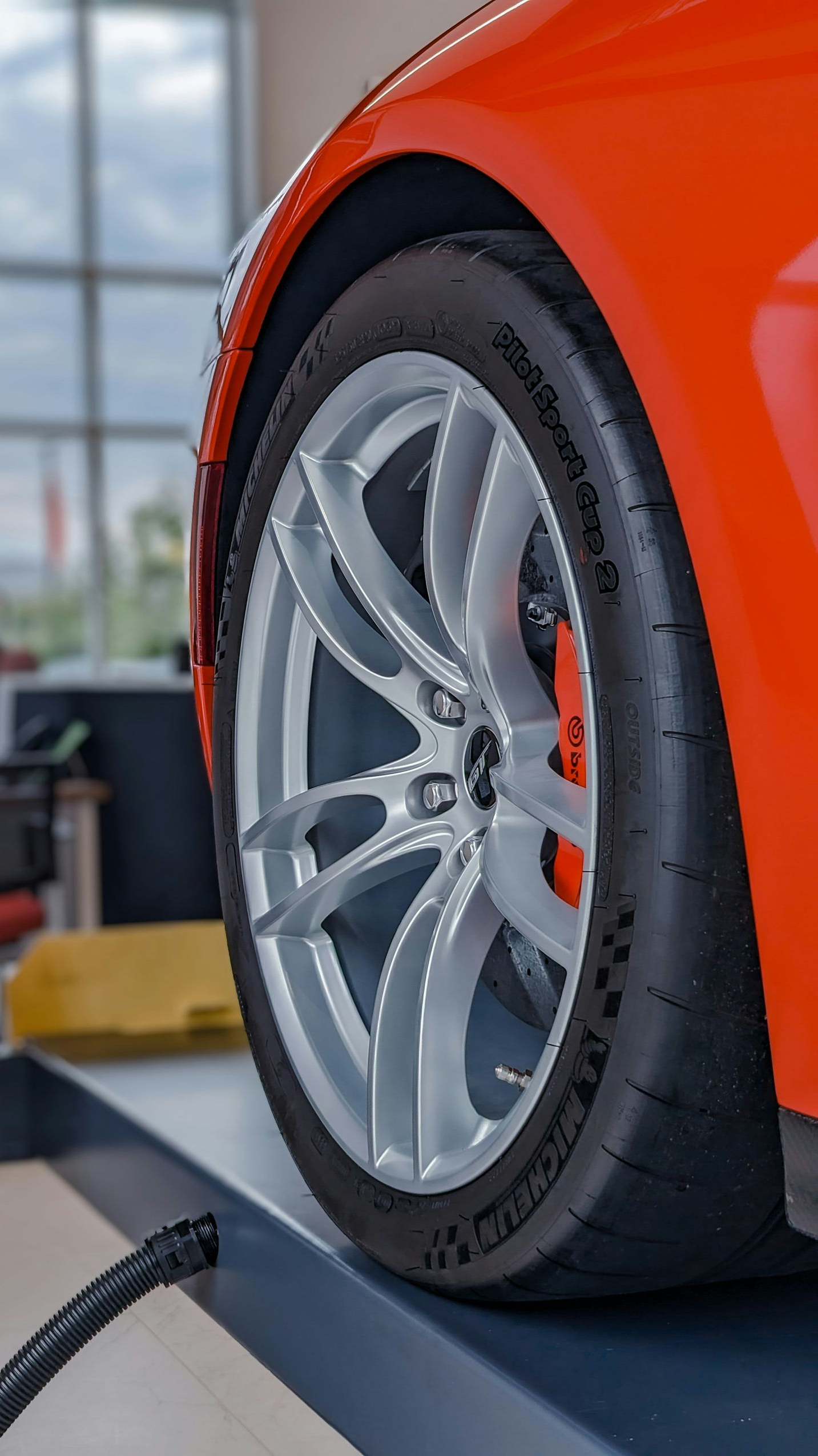 A bright orange car on a mechanic’s lift, showcasing a silver alloy wheel and high-performance tire in a shop.