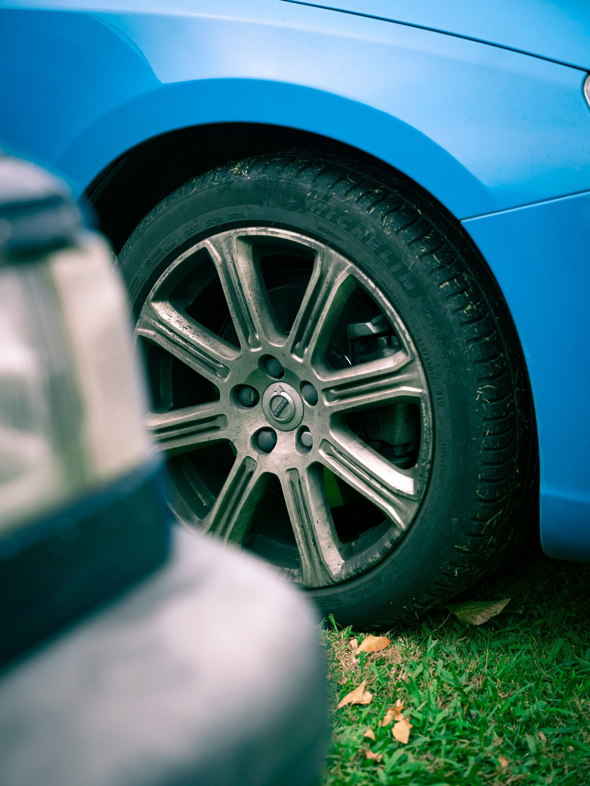 A bright blue car with a multi-spoke metallic wheel parked on grass, partially obscured by a vehicle in the foreground.