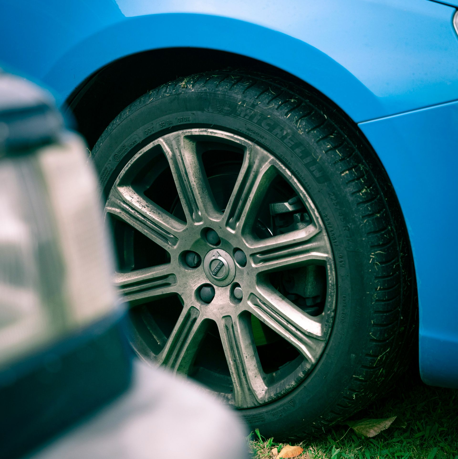 A bright blue car with a multi-spoke metallic wheel parked on grass, partially obscured by a vehicle in the foreground.