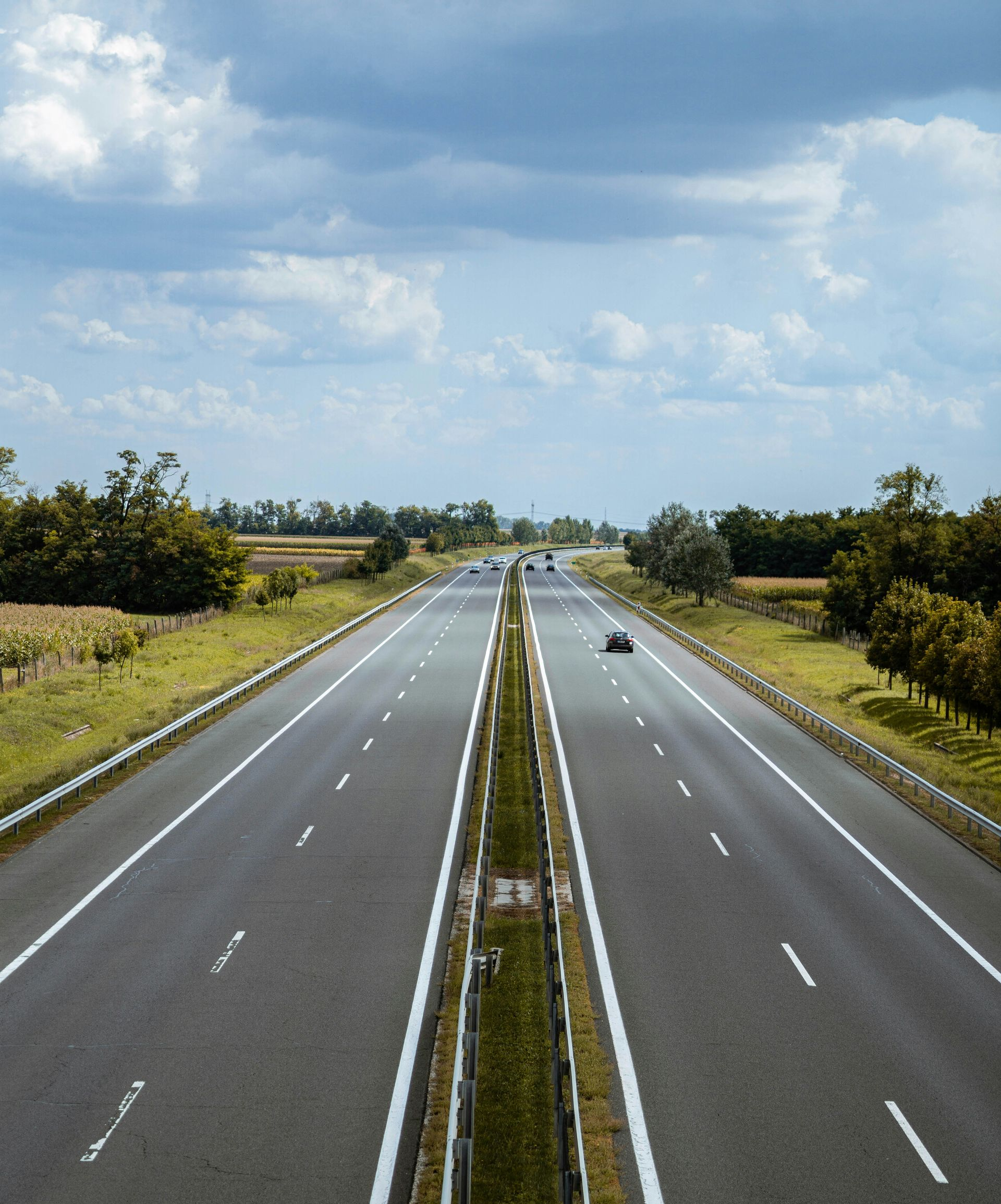 Empty multi-lane highway, gray asphalt, divided by a green median. A single car drives towards the horizon under a cloudy sky.