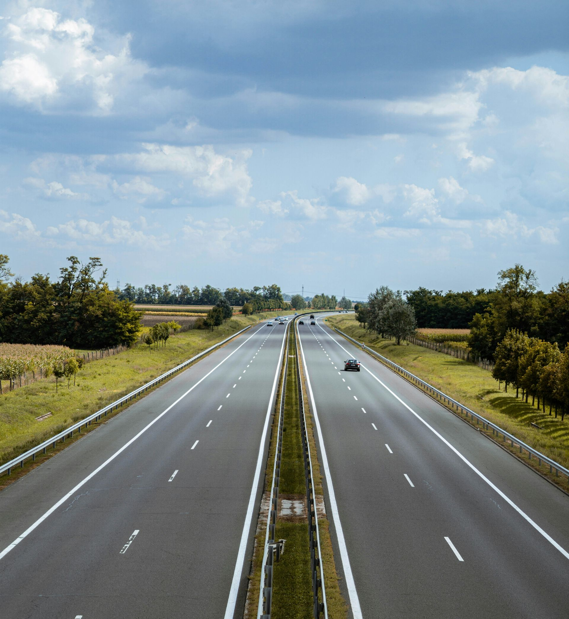 Empty multi-lane highway, gray asphalt, divided by a green median. A single car drives towards the horizon under a cloudy sky.
