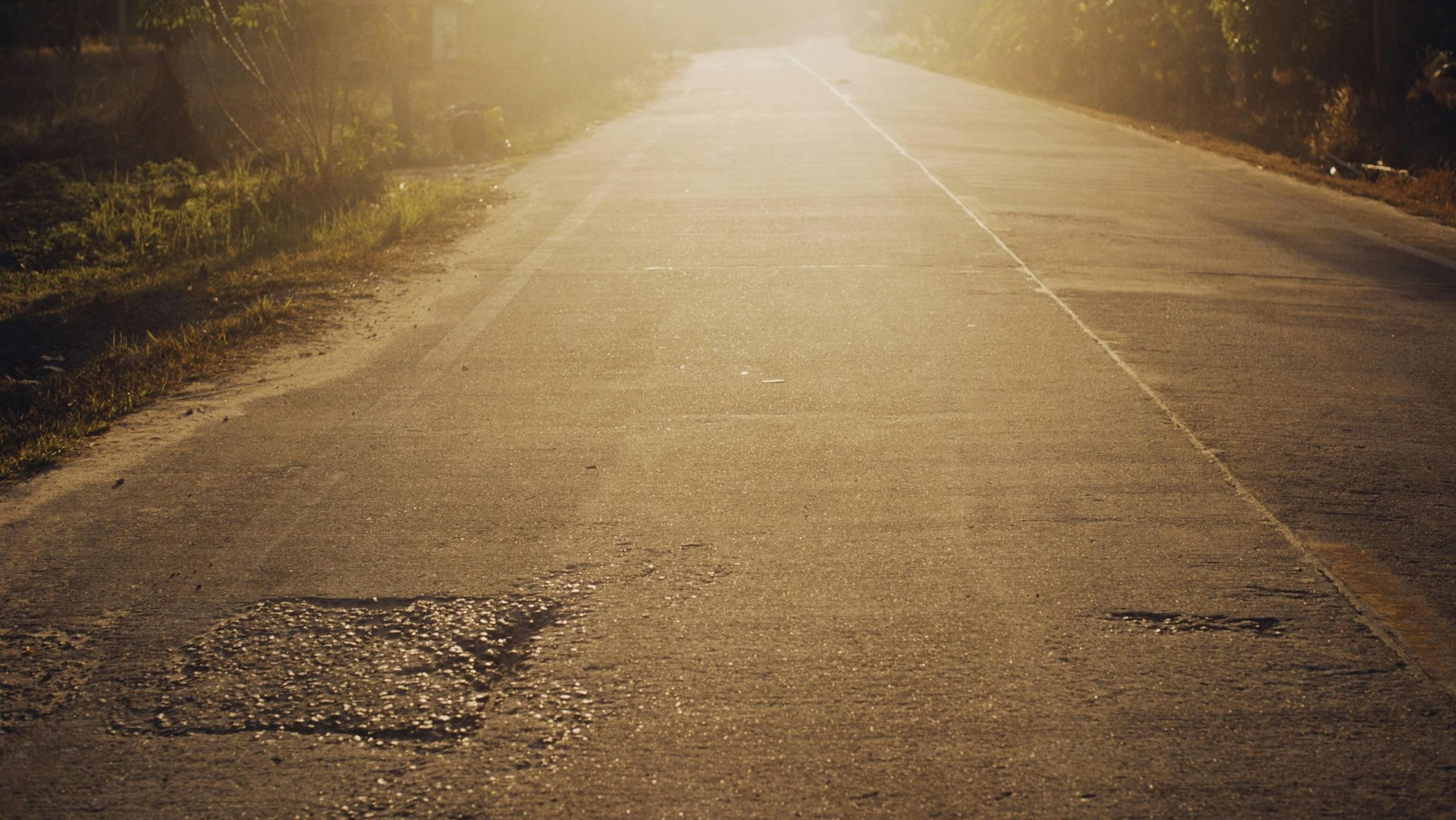 A rural asphalt road stretches into the distance, illuminated by a warm, hazy sunset filtering through overhanging trees.