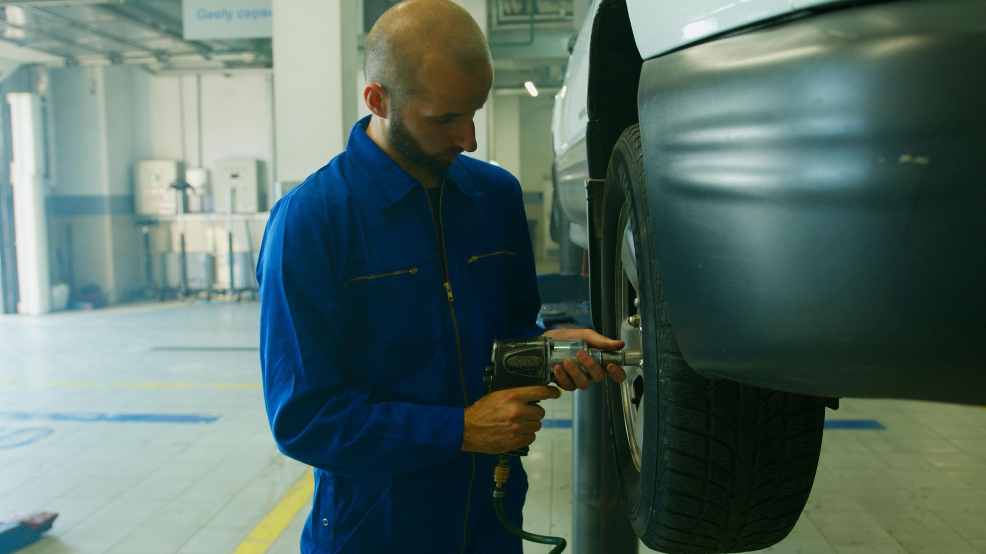 Mechanic in blue coveralls using a power tool to remove a tire in a garage.