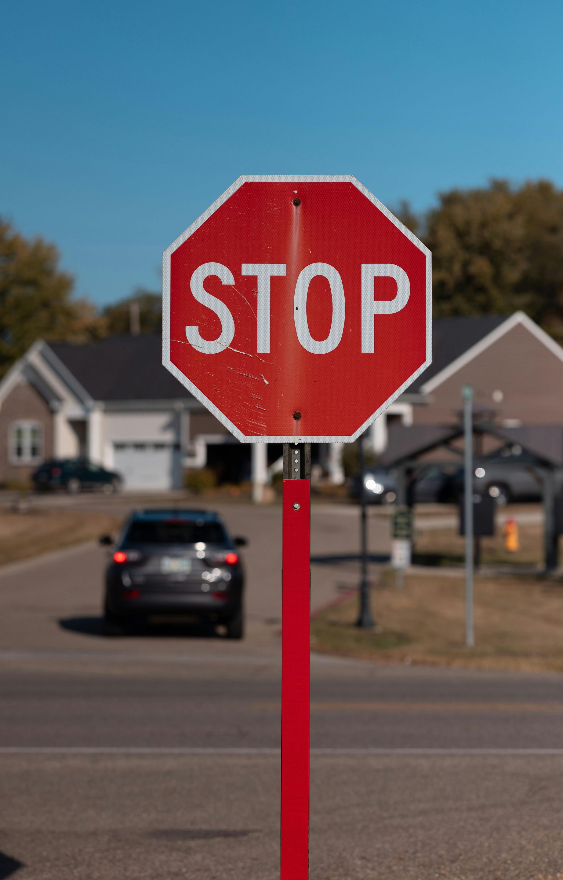 Red stop sign on a red pole in front of a street with a car and houses on a sunny day.